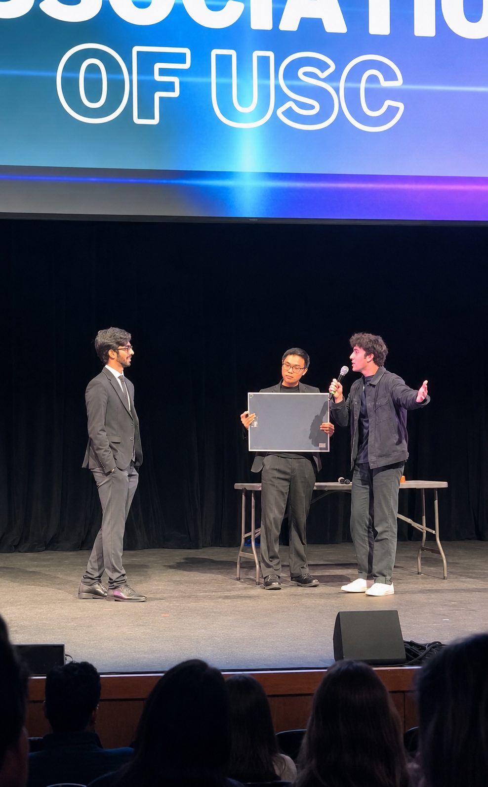 Tyler performing on stage at a USC Magic Association show, mic in hand, with two audience volunteers holding a board.