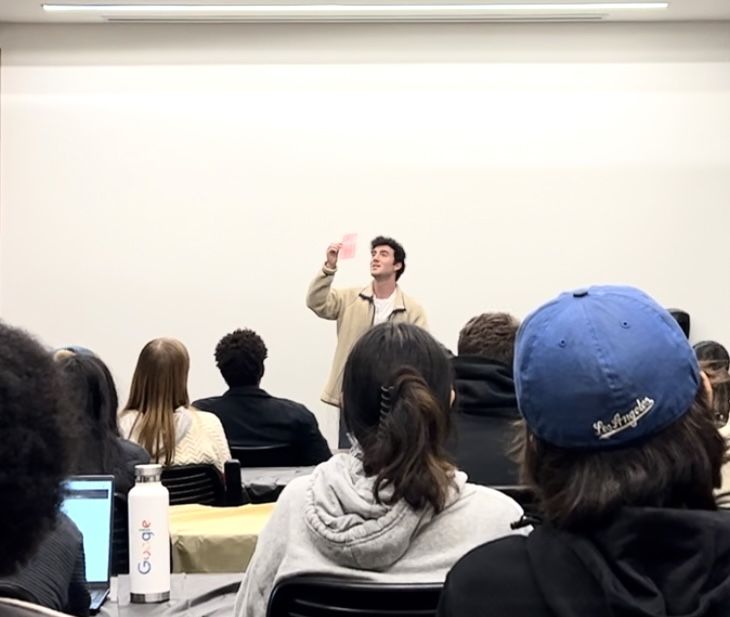 Tyler Gibgot holding a playing card above his head while lecturing to a seated class.