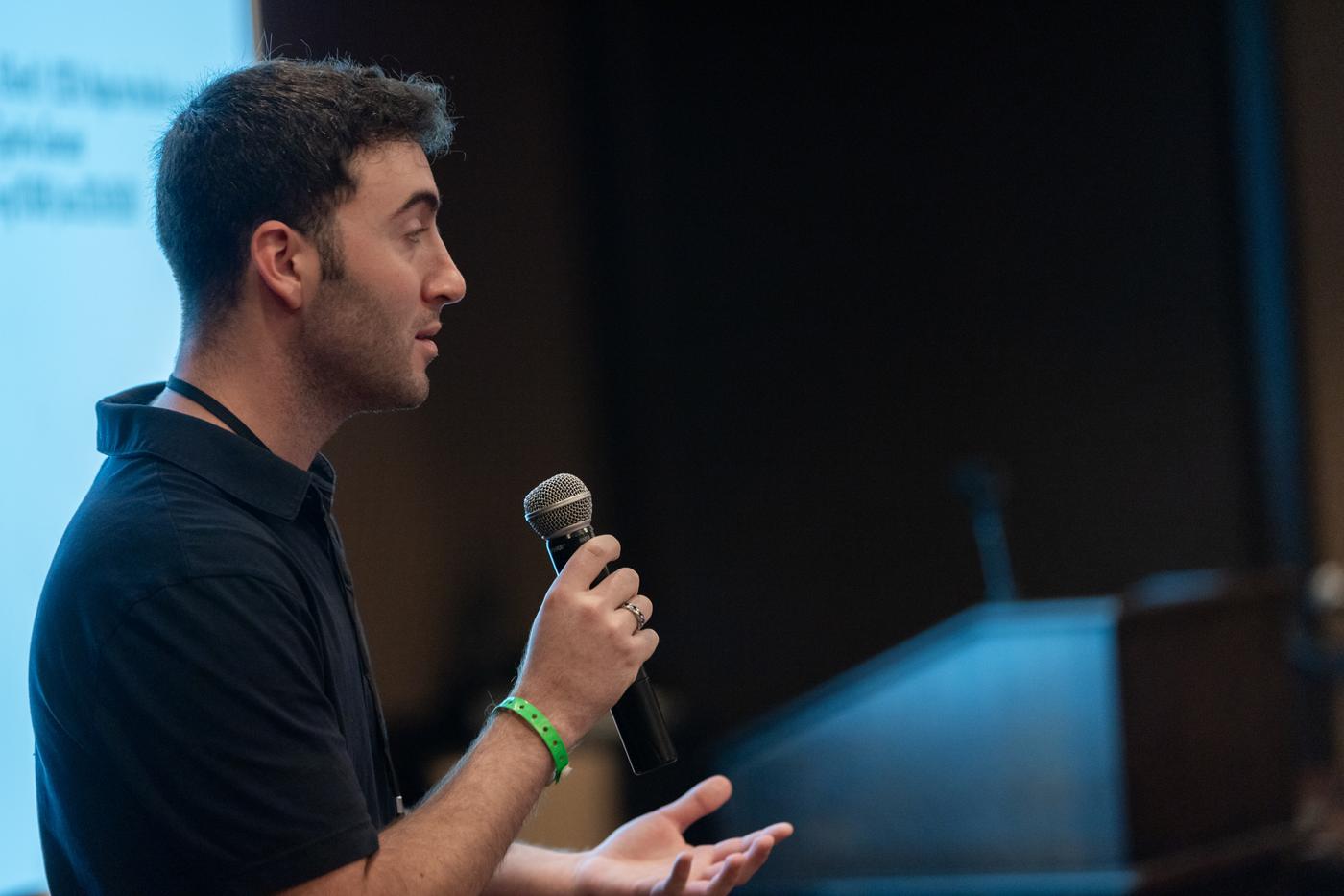 Tyler in profile holding a microphone, mid-sentence, presenting beside a projection screen.