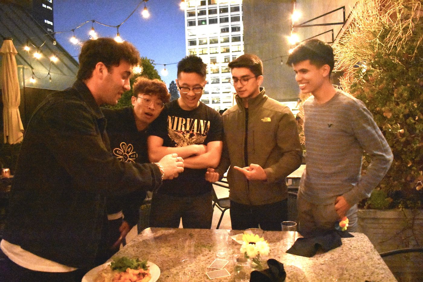 Five guests leaning in around a rooftop table, reacting as Tyler Gibgot finishes a close-up card trick.